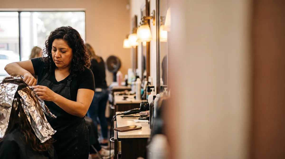 A salon owner foils a color treatment while her phone rests on the station, the AI making her win-back calls